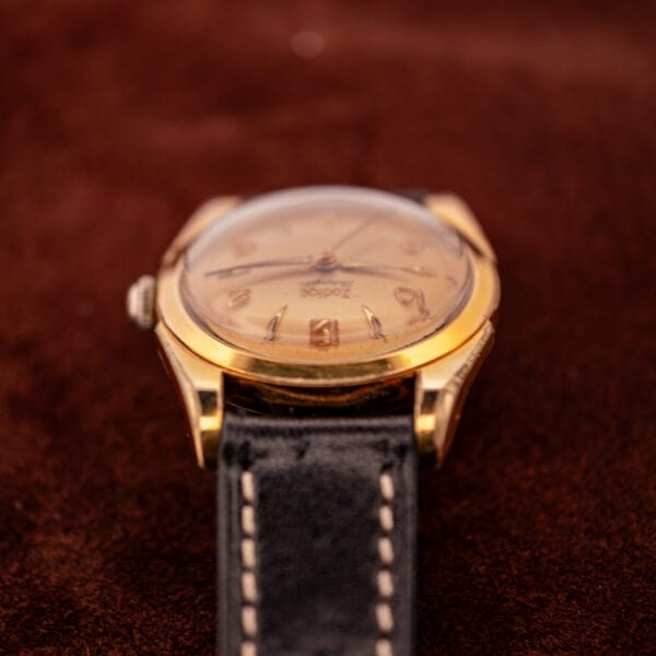 A Zodiac Rotographic Ref. 656 Tropic Dial Automatic Cal. 1381 wristwatch with a gold case and black leather band rests on a brown surface, viewed from a low angle.
