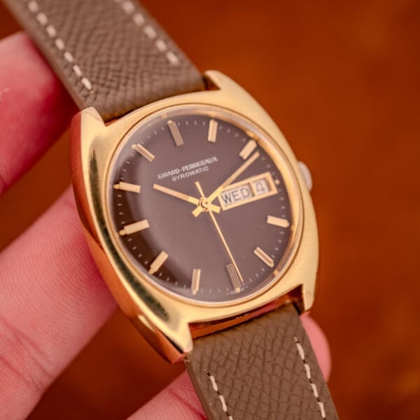 Hand-held wrist-scale photograph of the GP Gyromatic with brown dial, gold electroplate cushion case, and grey calfskin strap with contrast stitching.
