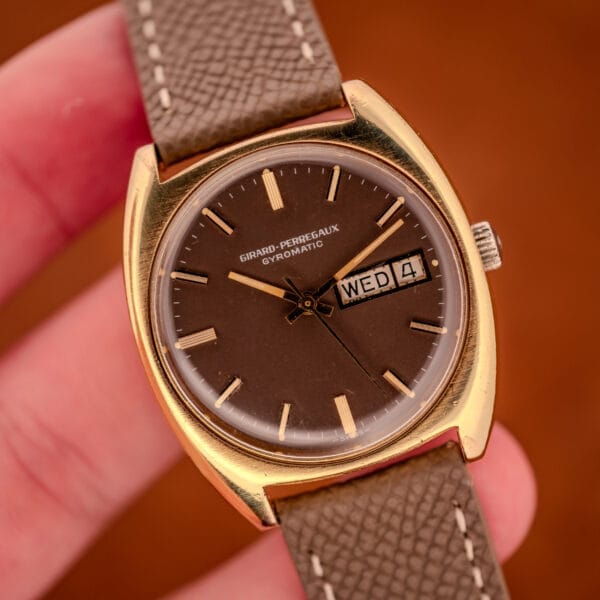Hand-held photograph of the Girard Perregaux Gyromatic 9276 HA showing the cushion case in scale, brown dial, and grey leather strap.
