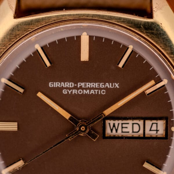 Macro of the Girard Perregaux Gyromatic chocolate brown dial showing the printed brand signature, applied gold ribbed batons, and day/date window.