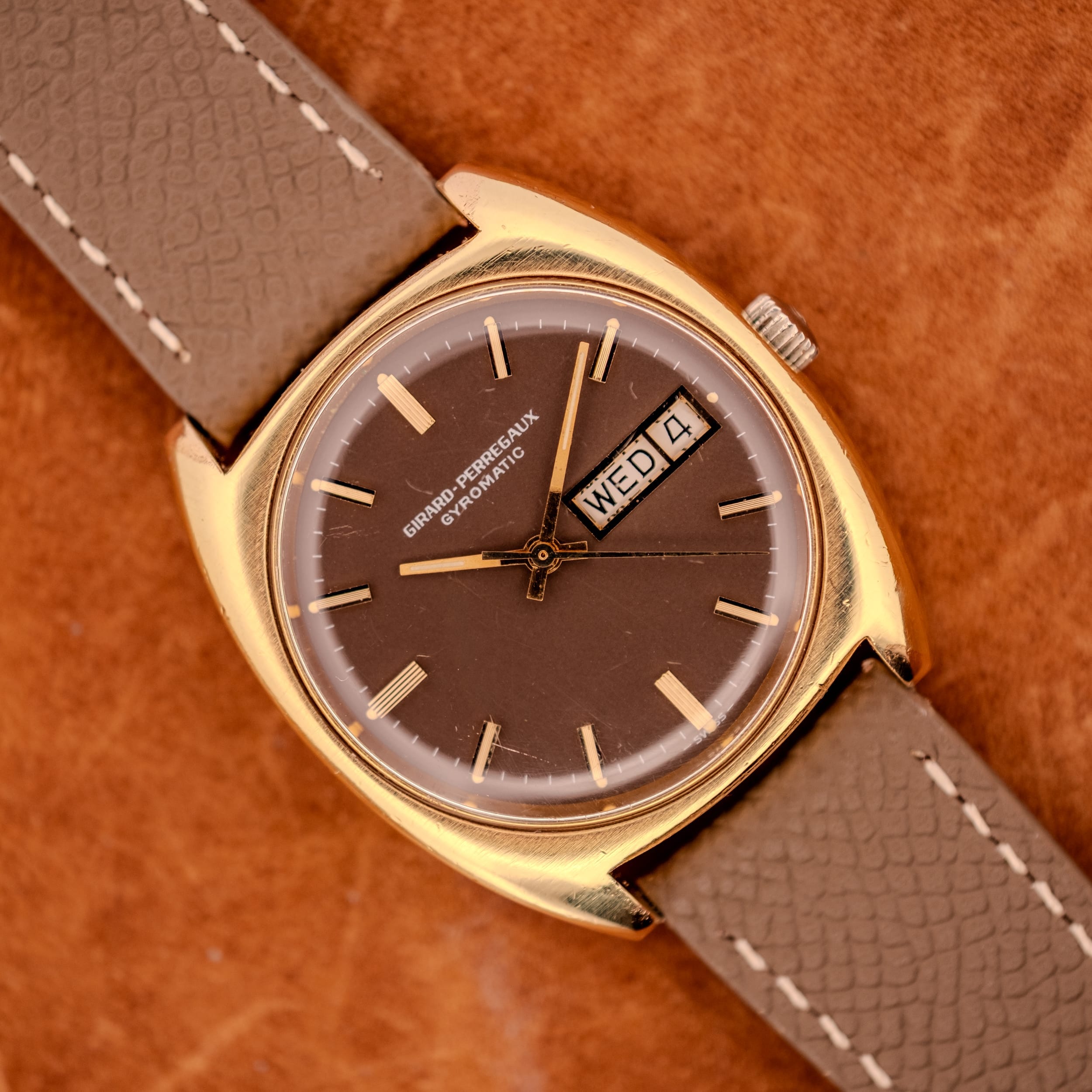 Girard Perregaux Gyromatic Ref. 9276 HA Day/Date Cal. 480-369 gold wristwatch with brown face, taupe strap, and day-date display, shown on a brown textured surface. Girard Perregaux Gyromatic Ref. 9276 HA front view, gold electroplate cushion case and chocolate brown dial with day/date at three.
