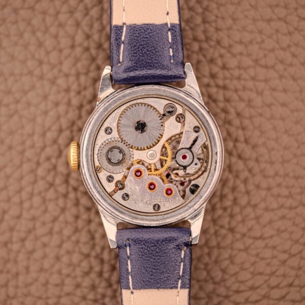 The Movado Manual Wind Sub-Seconds 14K Rose Gold Cap Sector Dial Ref. 13309, featuring an exposed mechanical movement and blue-beige strap, rests on a textured brown surface, with 14K rose gold accents adding elegance to its design.