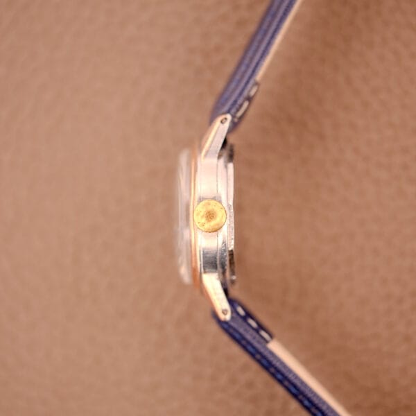 Side view of the Movado Manual Wind Sub-Seconds 14K Rose Gold Cap Sector Dial Ref. 13309 with a gold crown, blue strap, and a textured brown background.