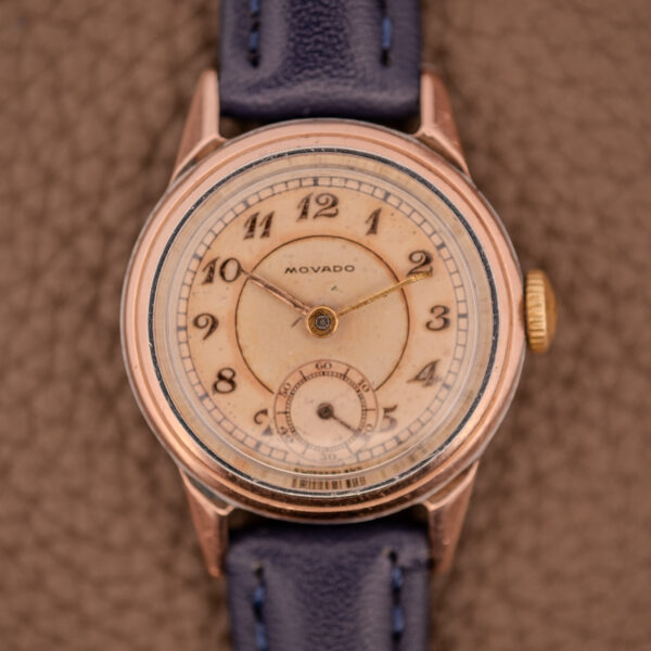 Movado Manual Wind Sub-Seconds Ref. 13309 with a 14K rose gold cap sector dial and blue leather strap, displayed on a textured brown surface.