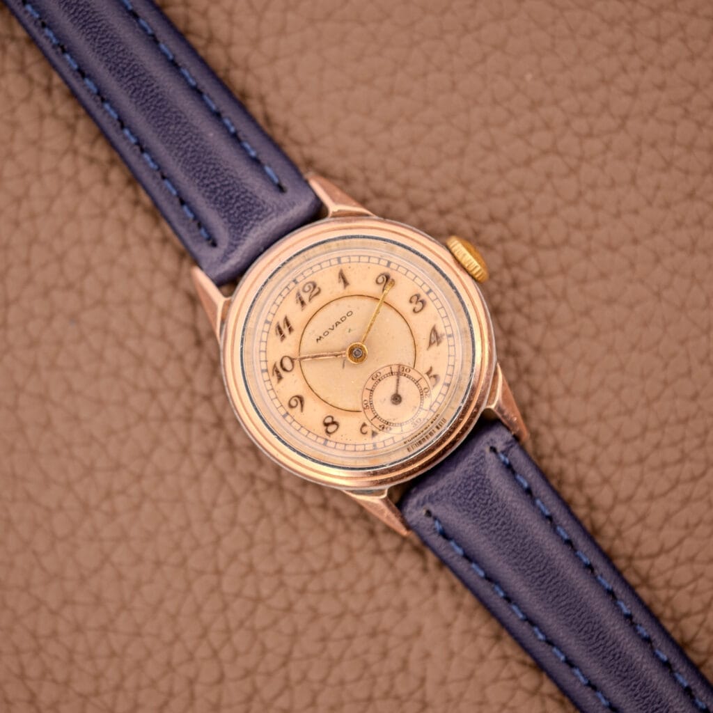 The Movado Manual Wind Sub-Seconds 14K Rose Gold Cap Sector Dial Ref. 13309 with a blue leather strap rests on a textured brown surface.