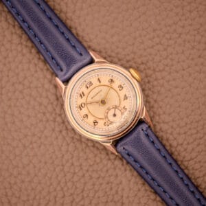 The Movado Manual Wind Sub-Seconds 14K Rose Gold Cap Sector Dial Ref. 13309 with a blue leather strap rests on a textured brown surface.