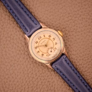 The Movado Manual Wind Sub-Seconds 14K Rose Gold Cap Sector Dial Ref. 13309 with a blue leather strap rests on a textured brown surface.