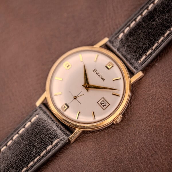 A Bulova Manual Wind Date 10K RGP Cal. 11ALD c.1968 wristwatch rests on a brown surface, featuring a gold case, black leather strap, and displaying the date as the 22nd.