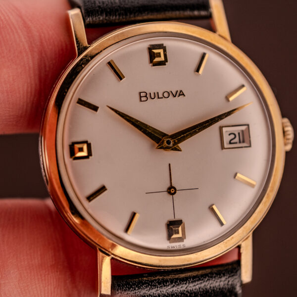 Close-up of a Bulova Manual Wind Date 10K RGP Cal. 11ALD c.1968 wristwatch with a white face, gold details, black strap, date display, and classic manual wind movement.