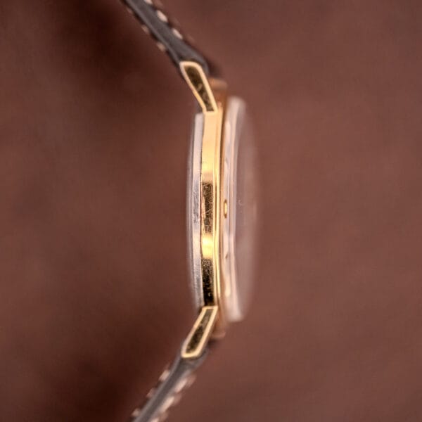 Close-up side view of a Bulova Manual Wind Date 10K RGP Cal. 11ALD c.1968, featuring a brown leather strap and gold case, set against a matching brown background.