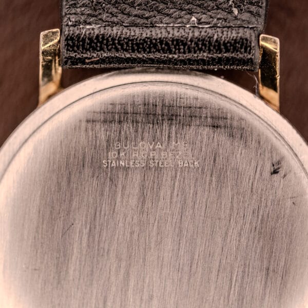 Close-up of a Bulova Manual Wind Date 10K RGP Cal. 11ALD c.1968, showing the engraved words STAINLESS STEEL BACK, 10K RGP case, and a black leather strap.