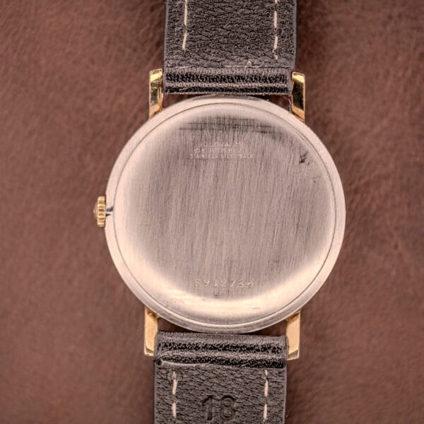 The back of a Bulova Manual Wind Date 10K RGP Cal. 11ALD c.1968 gold wristwatch with a black leather strap, marked 10K RGP, resting on a brown surface.