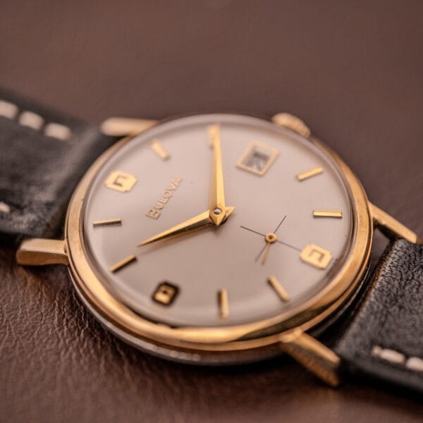 Close-up of a Bulova Manual Wind Date 10K RGP wristwatch c.1968 with Cal. 11ALD movement and black leather strap on a brown surface.