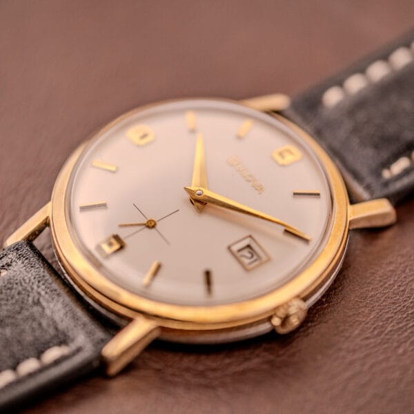 Close-up of a Bulova Manual Wind Date 10K RGP Cal. 11ALD c.1968 wristwatch with a black leather strap, displayed on a brown surface.
