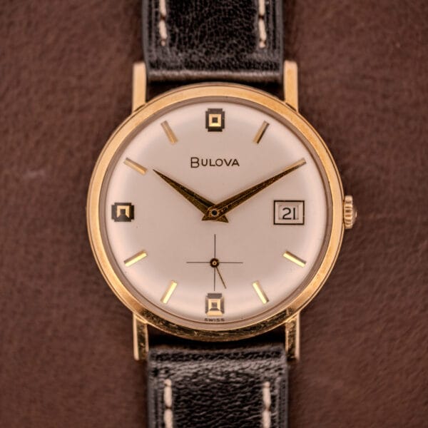 A Bulova Manual Wind Date 10K RGP Cal. 11ALD c.1968 wristwatch with a white dial, manual wind movement, black leather strap, and date display is shown on a brown surface.