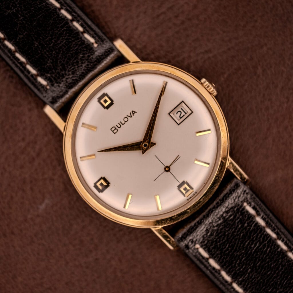 Bulova Manual Wind Date 10K RGP c.1968 wristwatch with Cal. 11ALD movement, white dial showing date “21,” and black leather strap.