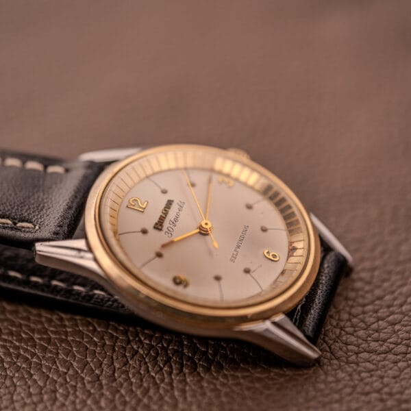 A Bulova 30 Jewels Selfwinding Unishell Case wristwatch (10K GF bezel, Cal. 10BZAC, c.1961) with a black leather strap rests on a brown textured surface.