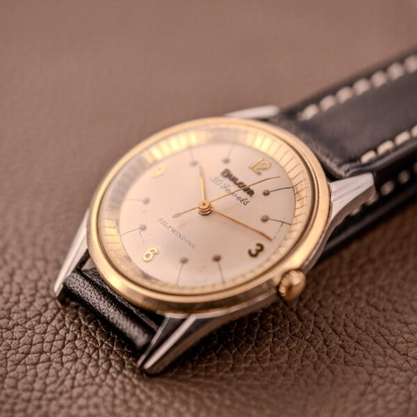The Bulova 30 Jewels Selfwinding Unishell Case wristwatch (Cal. 10BZAC, 10K GF bezel, c.1961) with a black leather strap rests on a brown textured surface.