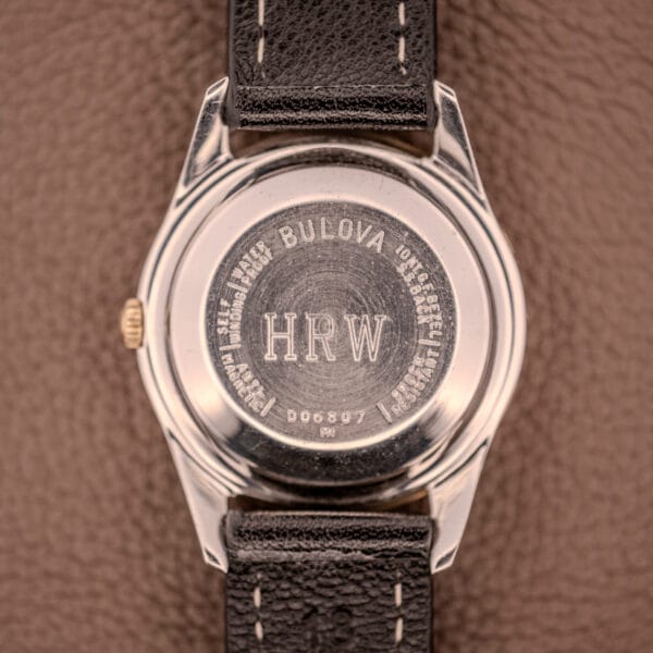 The back of a Bulova 30 Jewels Selfwinding Unishell Case 10K GF Bezel Cal. 10BZAC c.1961 wristwatch, engraved with HRW and serial number, rests on a brown textured surface.