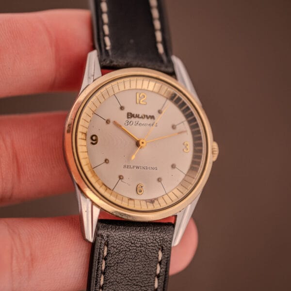 A hand holds a Bulova 30 Jewels Selfwinding Unishell Case watch (Cal. 10BZAC, c.1961) with a 10K GF bezel, white dial, black leather strap, and Arabic numerals.