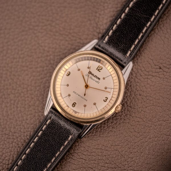Bulova 30 Jewels Selfwinding, Cal. 10BZAC (c.1961) with a Unishell case and 10K GF bezel, features a black leather strap; this vintage classic exudes timeless elegance on a textured brown background.
