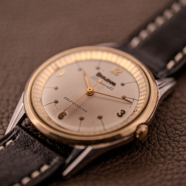 A Bulova 30 Jewels Selfwinding Unishell Case wristwatch, Cal. 10BZAC c.1961, with a 10K GF bezel and classic white dial on a leather strap, is displayed on a brown textured surface.