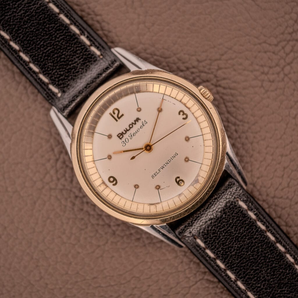 A Bulova 30 Jewels Selfwinding Unishell Case wristwatch (10K GF Bezel, Cal. 10BZAC, c.1961) with a black leather strap rests on a brown textured surface, displaying vintage elegance and automatic movement.