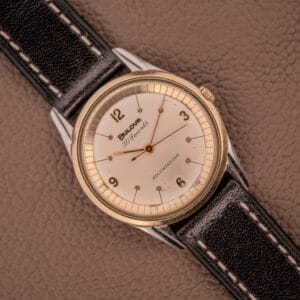A Bulova 30 Jewels Selfwinding Unishell Case wristwatch (10K GF Bezel, Cal. 10BZAC, c.1961) with a black leather strap rests on a brown textured surface, displaying vintage elegance and automatic movement.