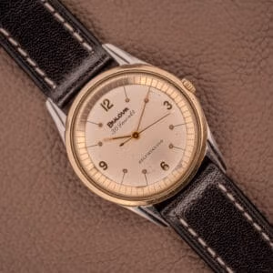 A Bulova 30 Jewels Selfwinding Unishell Case wristwatch (10K GF Bezel, Cal. 10BZAC, c.1961) with a black leather strap rests on a brown textured surface, displaying vintage elegance and automatic movement.