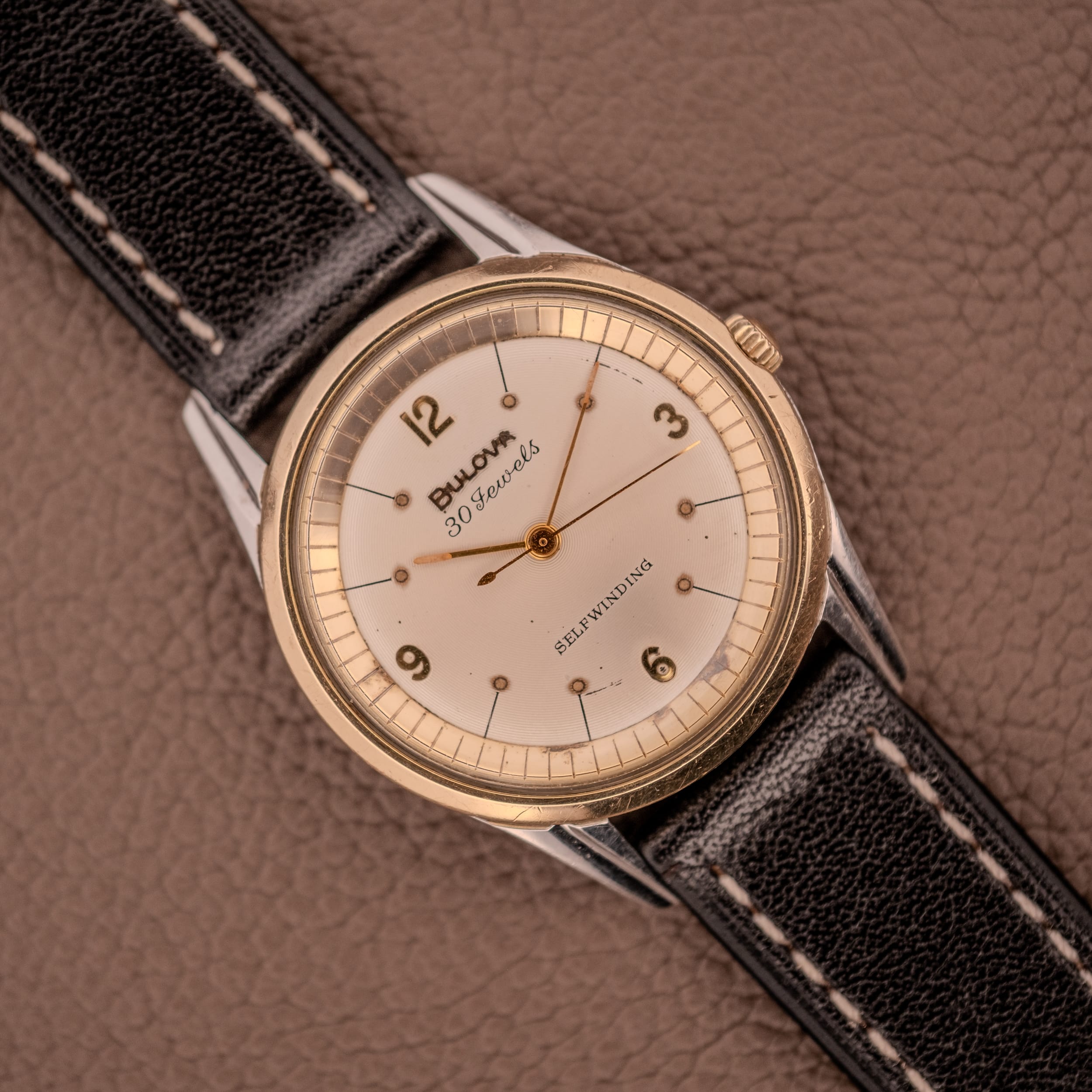A Bulova 30 Jewels Selfwinding Unishell Case wristwatch (10K GF Bezel, Cal. 10BZAC, c.1961) with a black leather strap rests on a brown textured surface, displaying vintage elegance and automatic movement. A Bulova 30 Jewels Selfwinding Unishell Case wristwatch (10K GF Bezel, Cal. 10BZAC, c.1961) with a black leather strap rests on a brown textured surface, displaying vintage elegance and automatic movement.