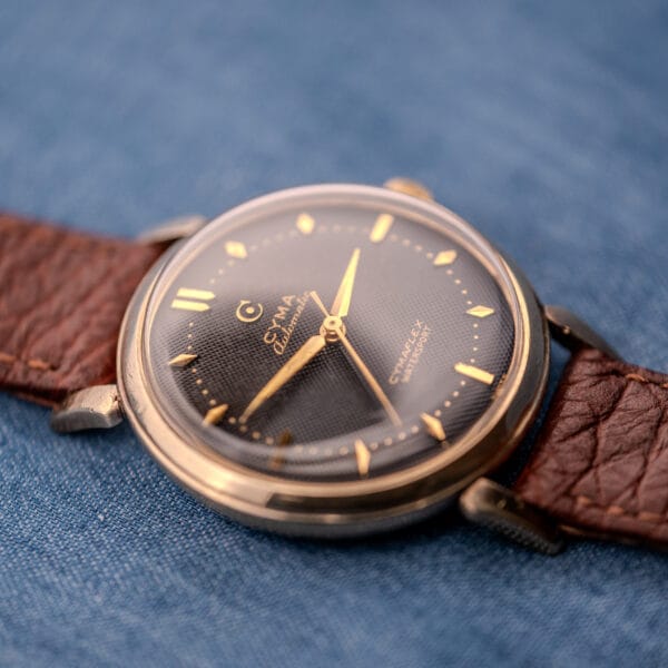 A Cyma Cymaflex Watersport Automatic wristwatch, featuring a Black Honeycomb Dial (Cal. R.420) and brown leather strap, displayed on blue fabric.