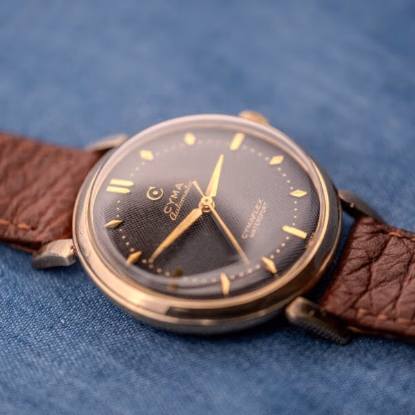 Three-quarter shot of the 1950s Cyma Watersport, slim gold-tone case and black waffle dial on blue cloth.