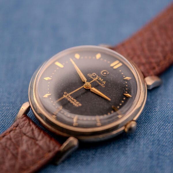 The Cyma Cymaflex Watersport Automatic, featuring a black honeycomb dial (Cal. R.420) and a brown leather strap, is displayed on a blue fabric background.