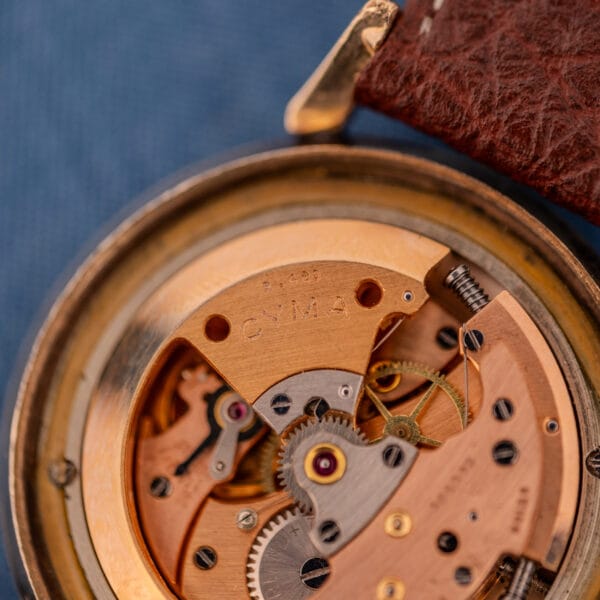 Close-up of a Cyma Cymaflex Watersport Automatic watch featuring a black honeycomb dial, Cal. R.420 movement with visible gears, and a brown leather strap.