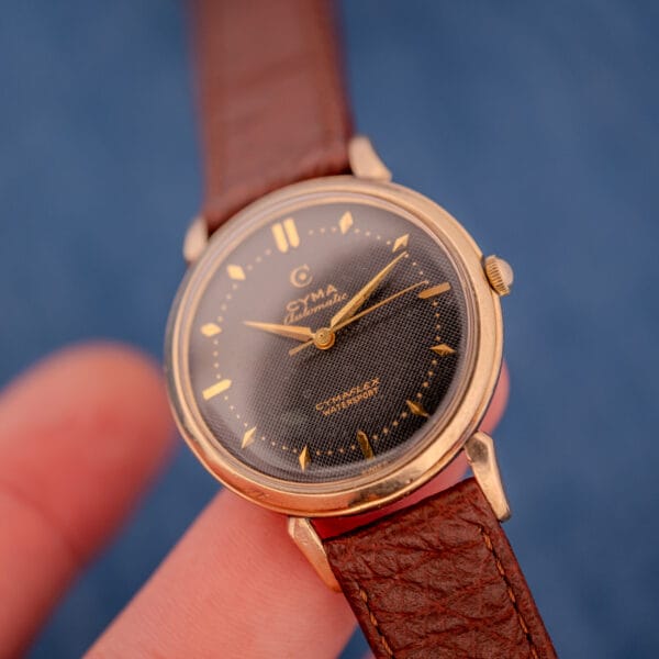 A close-up of the Cyma Cymaflex Watersport Automatic with Cal. R.420, featuring a black honeycomb dial, gold accents, and a brown leather strap against a blue background.