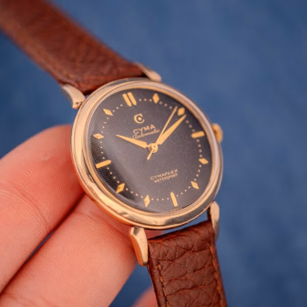 1950s Cyma Watersport held in hand, black honeycomb dial and gold dauphine hands catching natural light.