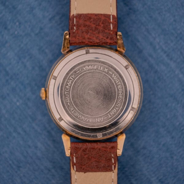 The back of a Cyma Cymaflex Watersport Automatic Black Honeycomb Dial Cal. R.420 watch with a brown leather strap, featuring engraved text, displayed on a blue fabric background.