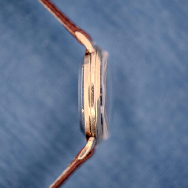 Side view of the Cyma Cymaflex Watersport Automatic Black Honeycomb Dial Cal. R.420 gold wristwatch with brown leather strap, set against a blue fabric background.