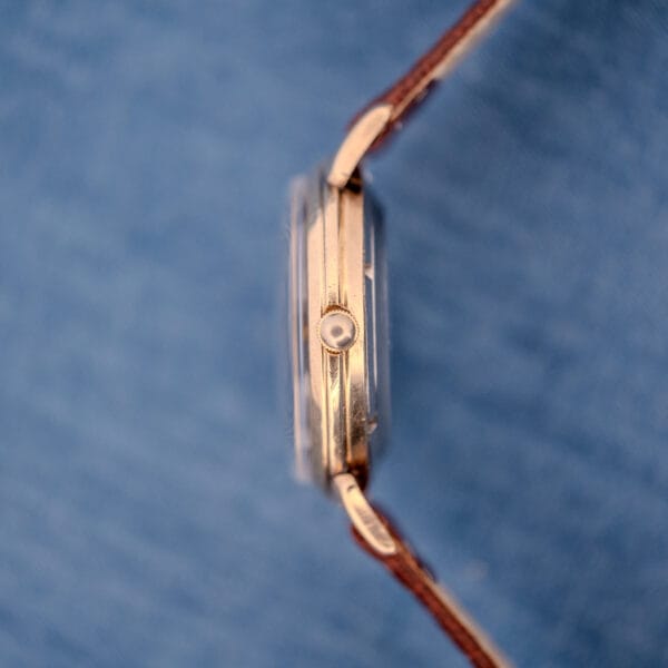 Side view of a gold Cyma Cymaflex Watersport Automatic Black Honeycomb Dial Cal. R.420 wristwatch with a brown leather strap, set against a blue textured background.