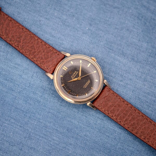 A Cyma Cymaflex Watersport Automatic wristwatch with a black honeycomb dial (Cal. R.420) and brown leather strap rests on a blue denim background.