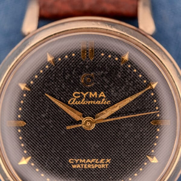 Close-up of a Cyma Cymaflex Watersport Automatic wristwatch featuring gold hands and markers on a textured black honeycomb dial, powered by Cal. R.420.