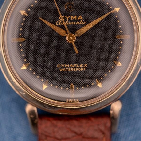 Close-up of a Cyma Cymaflex Watersport Automatic watch featuring gold hands, black honeycomb dial (Cal. R.420), and a brown leather strap.