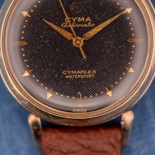 Cyma Watersport dial detail showing CYMAFLEX WATERSPORT signature, SWISS print, gold C-with-arrow logo, and honeycomb texture.