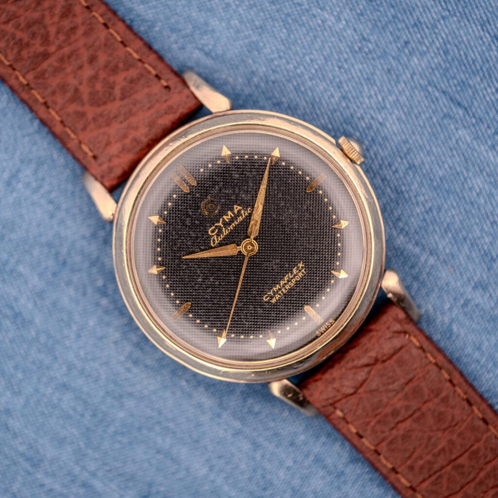 The Cyma Cymaflex Watersport Automatic Black Honeycomb Dial Cal. R.420, a vintage gold wristwatch with a brown leather strap, rests on blue denim fabric.