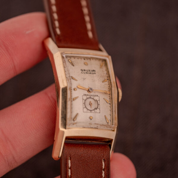 A hand holding a Gruen Curvex Precision "Pyramid Crystal" 10K GF Ref. 370-601 wristwatch, featuring a brown leather strap and rectangular face.