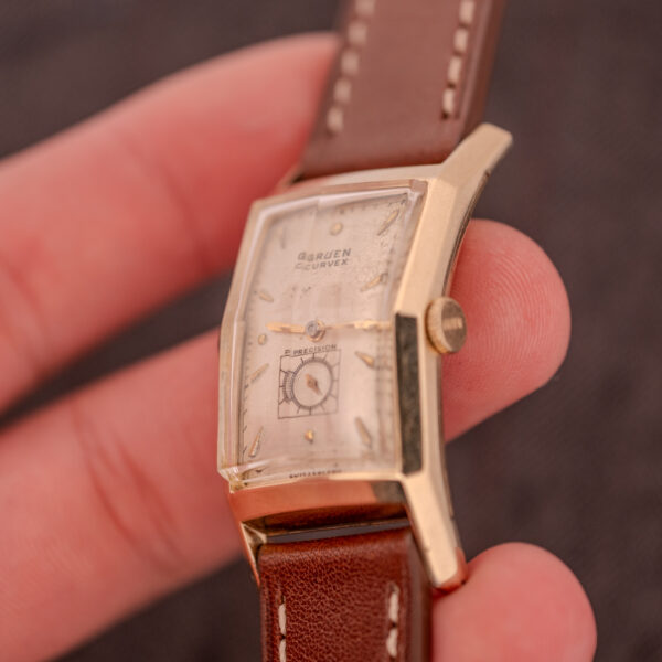 A hand holds a Gruen Curvex Precision "Pyramid Crystal" 10K GF Ref. 370-601 wristwatch with a rectangular gold case and brown leather strap.