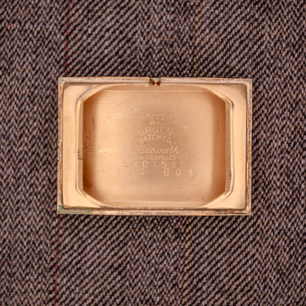 A rectangular gold Gruen Curvex Precision "Pyramid Crystal" 10K GF Ref. 370-601 watch case back with engraved text, marked 10K GF, is placed on brown patterned fabric.