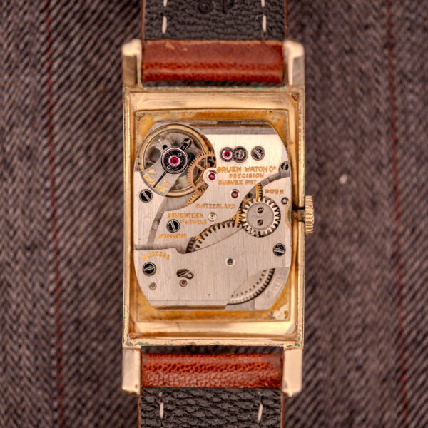 Close-up of a Gruen Curvex Precision "Pyramid Crystal" 10K GF Ref. 370-601 watch movement with visible gears, set against a striped fabric background.