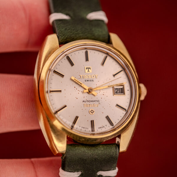 A Tissot Seastar Automatic Date Ref. 44660 Cal. 786-2 from the 1970s, featuring a gold case, white dial, and green strap, is displayed against a bold red background.
