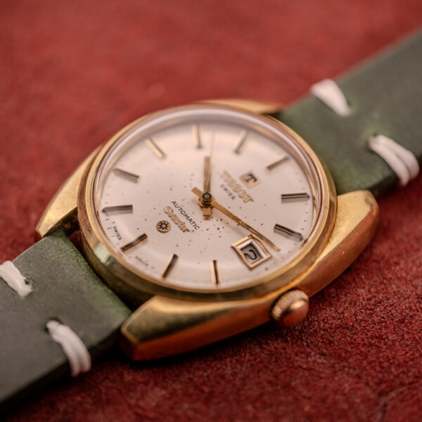 Close-up of a vintage Tissot Seastar Automatic Date Ref. 44660 Cal. 786-2 from the 1970s, featuring a gold case, white dial, and green leather strap, displayed on a red surface.